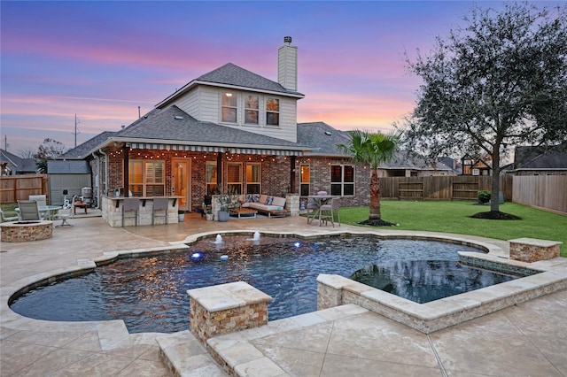 pool at dusk featuring an outdoor living space with a fire pit, exterior bar, a lawn, and a patio