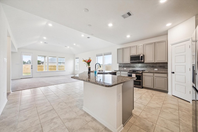 kitchen featuring light carpet, stainless steel appliances, dark stone counters, decorative backsplash, and a kitchen island with sink