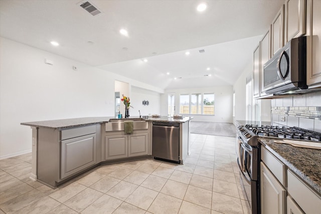 kitchen with stainless steel appliances, dark stone counters, an island with sink, sink, and vaulted ceiling