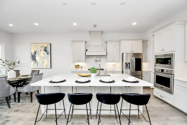 kitchen featuring custom exhaust hood, white cabinets, appliances with stainless steel finishes, and an island with sink