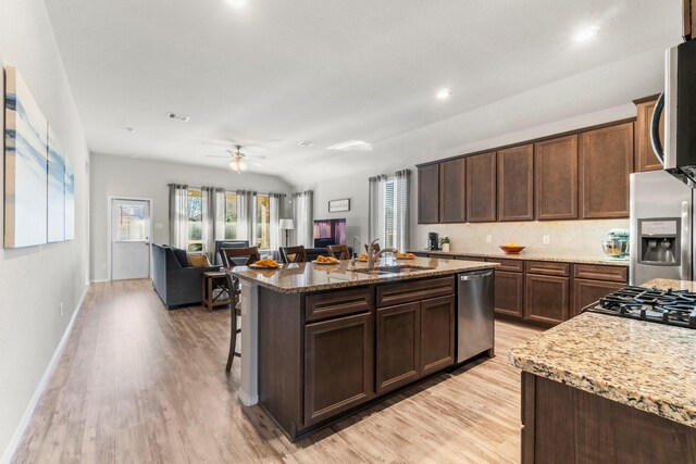 kitchen with stone counters, an island with sink, sink, stainless steel appliances, and light wood-type flooring