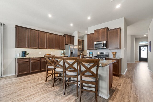 kitchen featuring a kitchen island with sink, stainless steel appliances, a kitchen breakfast bar, light stone counters, and decorative backsplash