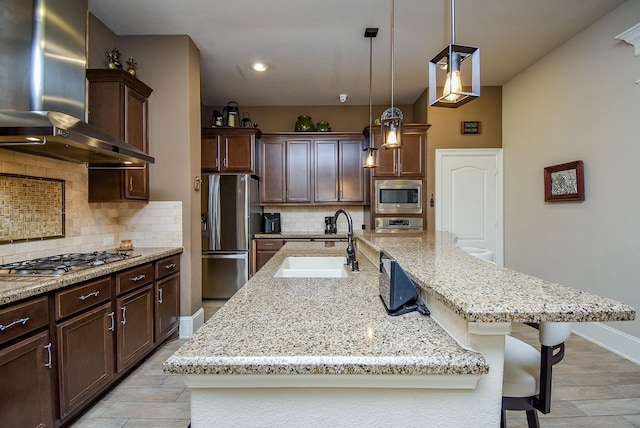 kitchen with wall chimney range hood, sink, stainless steel appliances, an island with sink, and decorative backsplash