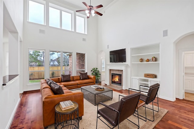 living room with a towering ceiling, wood-type flooring, built in features, and ceiling fan