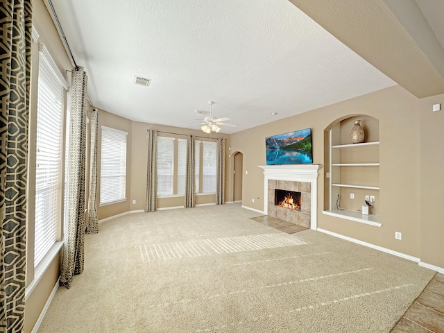 unfurnished living room with a tile fireplace, built in features, carpet floors, ceiling fan, and a textured ceiling