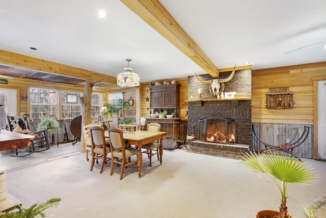 dining room featuring beamed ceiling, wooden walls, a brick fireplace, and concrete floors