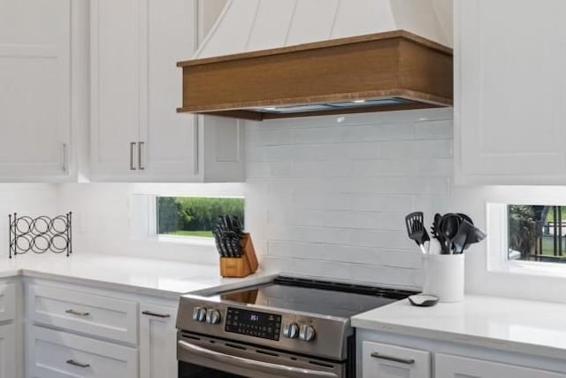 kitchen with stainless steel electric stove, custom range hood, white cabinets, light countertops, and tasteful backsplash