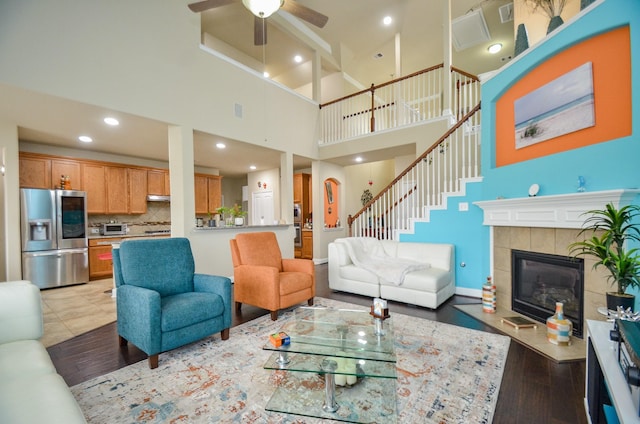 living room with a tile fireplace, light wood-type flooring, a towering ceiling, and ceiling fan