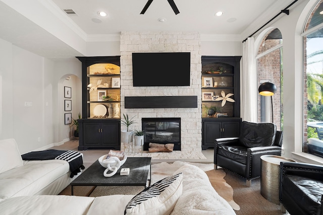 living room with built in features, crown molding, a stone fireplace, and a healthy amount of sunlight