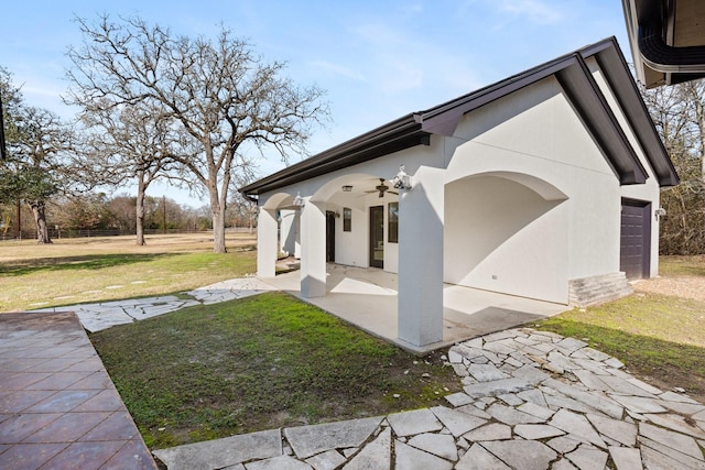 exterior space featuring a garage, a lawn, a patio, a ceiling fan, and stucco siding