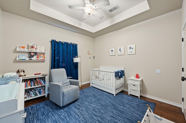 bedroom with ceiling fan, dark wood-type flooring, a raised ceiling, and a crib