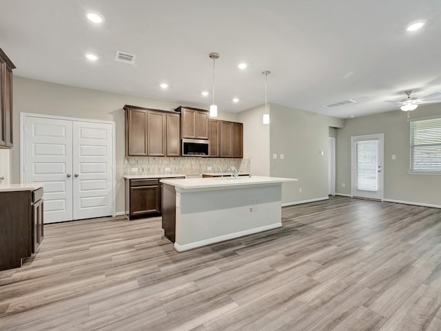 kitchen with ceiling fan, light hardwood / wood-style floors, decorative backsplash, hanging light fixtures, and a kitchen island with sink