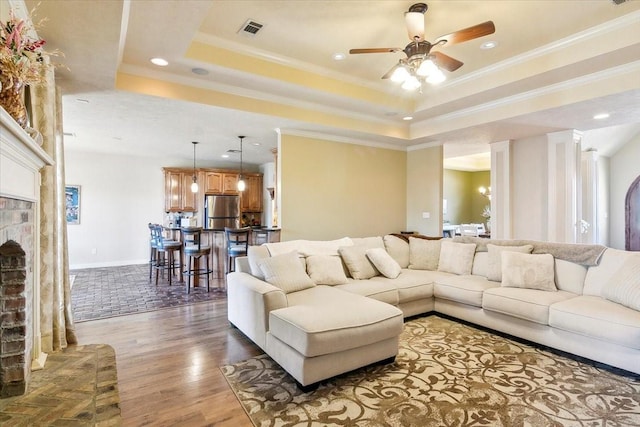 living room featuring a brick fireplace, dark hardwood / wood-style floors, a tray ceiling, ornamental molding, and ceiling fan with notable chandelier