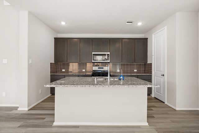 kitchen with tasteful backsplash, a center island with sink, dark brown cabinetry, light stone countertops, and appliances with stainless steel finishes