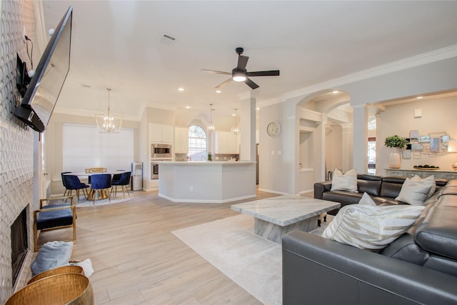 living room with light hardwood / wood-style flooring, a tiled fireplace, ornamental molding, ceiling fan with notable chandelier, and decorative columns