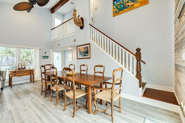 dining space with ceiling fan, beamed ceiling, a high ceiling, and light wood-type flooring