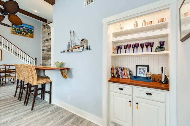 bar with wooden counters, light hardwood / wood-style flooring, beamed ceiling, ceiling fan, and white cabinets