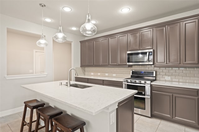 kitchen with sink, an island with sink, pendant lighting, stainless steel appliances, and light stone countertops