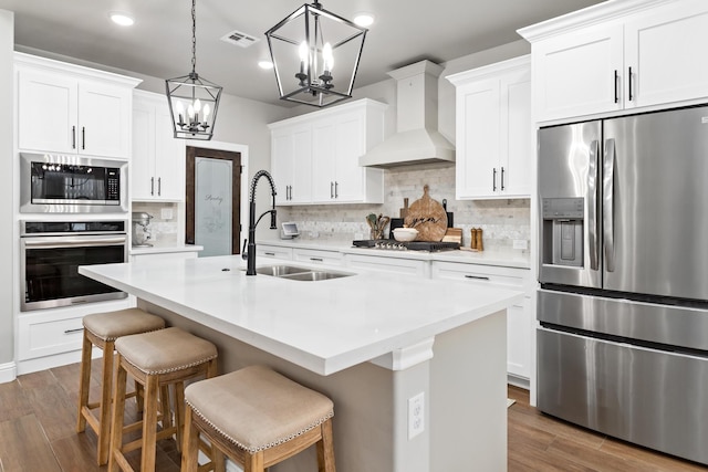 kitchen with custom exhaust hood, appliances with stainless steel finishes, white cabinets, sink, and a center island with sink