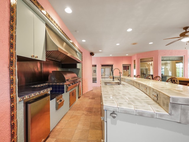 kitchen featuring island range hood, sink, white cabinets, tile countertops, and stainless steel fridge