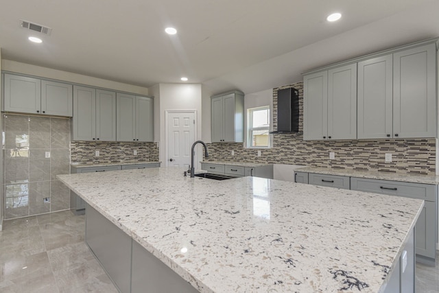 kitchen featuring sink, light stone counters, a center island with sink, wall chimney exhaust hood, and gray cabinets