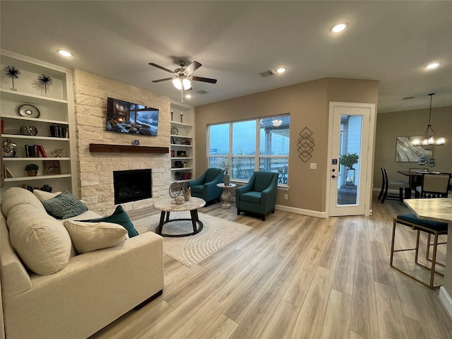 living room featuring built in shelves, a stone fireplace, ceiling fan with notable chandelier, and light hardwood / wood-style flooring