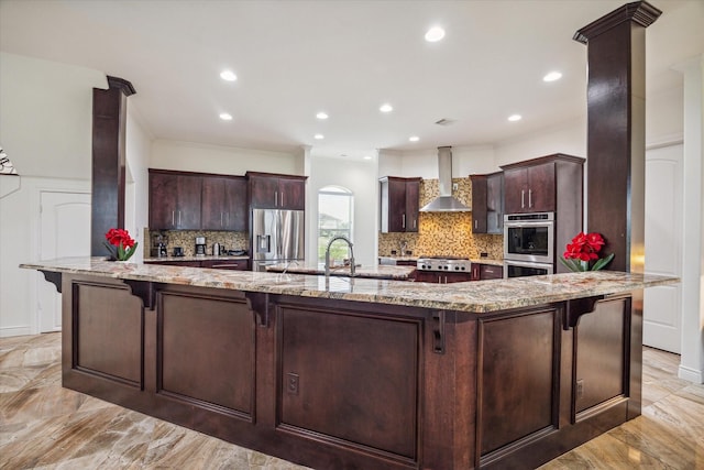 kitchen featuring wall chimney range hood, light stone counters, a kitchen bar, and stainless steel refrigerator with ice dispenser