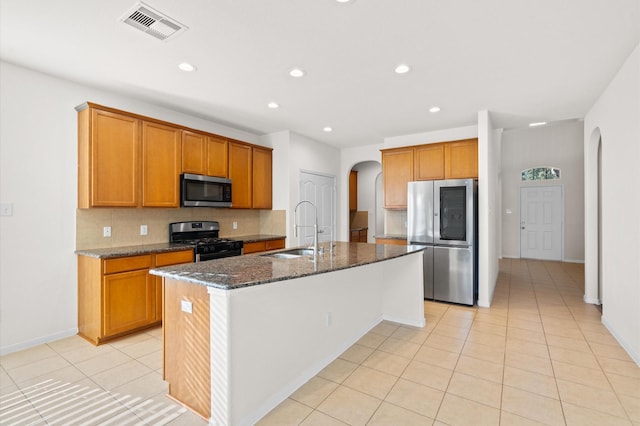 kitchen featuring appliances with stainless steel finishes, dark stone counters, sink, backsplash, and an island with sink