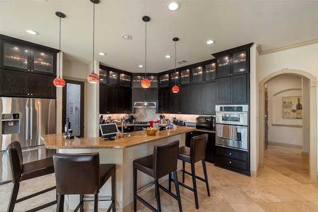 kitchen with arched walkways, stainless steel appliances, a kitchen island with sink, under cabinet range hood, and a kitchen bar