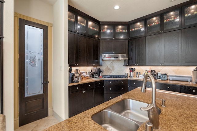 kitchen with glass insert cabinets, light stone counters, a sink, backsplash, and exhaust hood