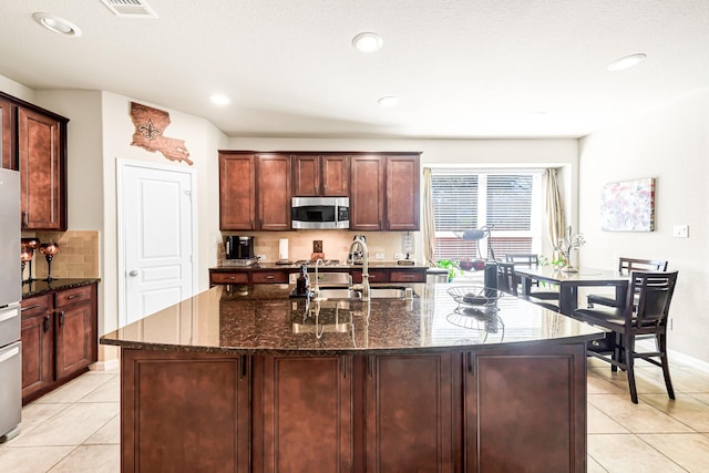 kitchen featuring tasteful backsplash, light tile patterned floors, dark stone counters, a kitchen island with sink, and stainless steel appliances