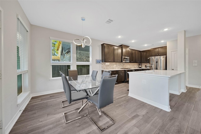 kitchen with appliances with stainless steel finishes, light wood-type flooring, decorative light fixtures, dark brown cabinets, and a center island with sink