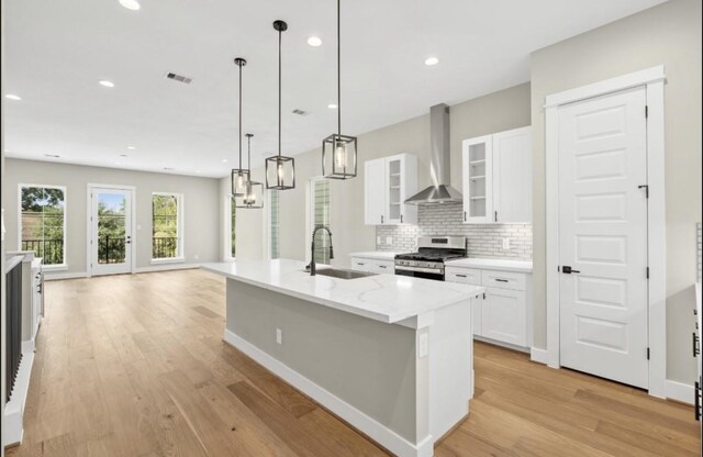 kitchen with stainless steel gas range, sink, wall chimney range hood, a kitchen island with sink, and white cabinets