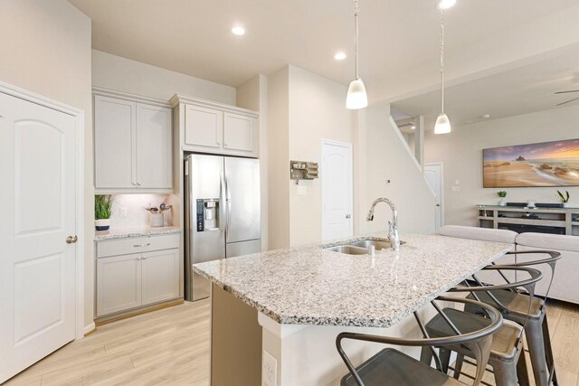 kitchen featuring sink, hanging light fixtures, a kitchen island with sink, stainless steel refrigerator with ice dispenser, and light stone countertops