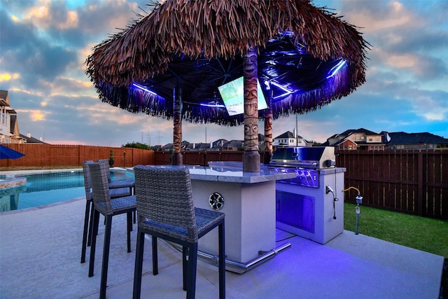 patio terrace at dusk featuring area for grilling, a fenced in pool, an outdoor bar, and an outdoor kitchen