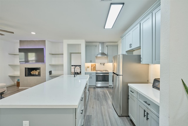 kitchen featuring appliances with stainless steel finishes, backsplash, light wood-type flooring, wall chimney range hood, and an island with sink