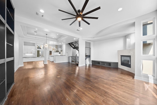 unfurnished living room featuring hardwood / wood-style flooring, a tray ceiling, a tiled fireplace, and ceiling fan with notable chandelier