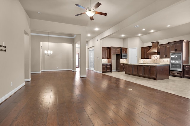 kitchen with appliances with stainless steel finishes, custom range hood, decorative backsplash, ceiling fan with notable chandelier, and a center island with sink