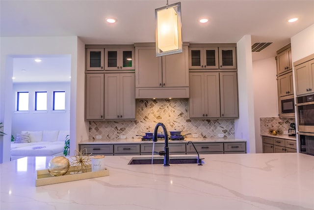 kitchen with sink, backsplash, stainless steel appliances, light stone counters, and decorative light fixtures