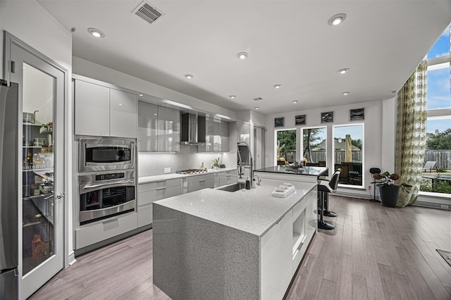 kitchen with light hardwood / wood-style flooring, sink, a kitchen island with sink, wall chimney range hood, and stainless steel appliances