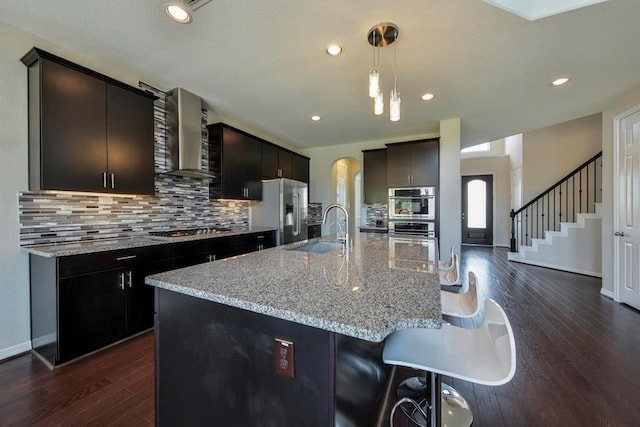 kitchen featuring dark hardwood / wood-style floors, sink, stainless steel appliances, a center island with sink, and wall chimney exhaust hood