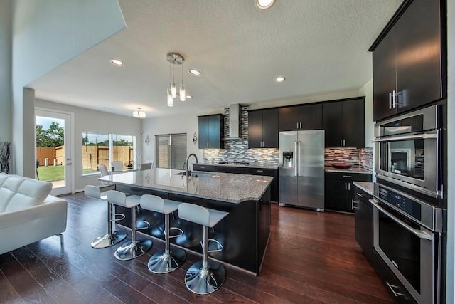 kitchen with wall chimney exhaust hood, sink, a center island with sink, appliances with stainless steel finishes, and pendant lighting