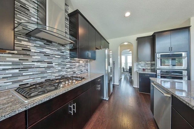 kitchen with appliances with stainless steel finishes, backsplash, light stone counters, dark hardwood / wood-style flooring, and wall chimney exhaust hood