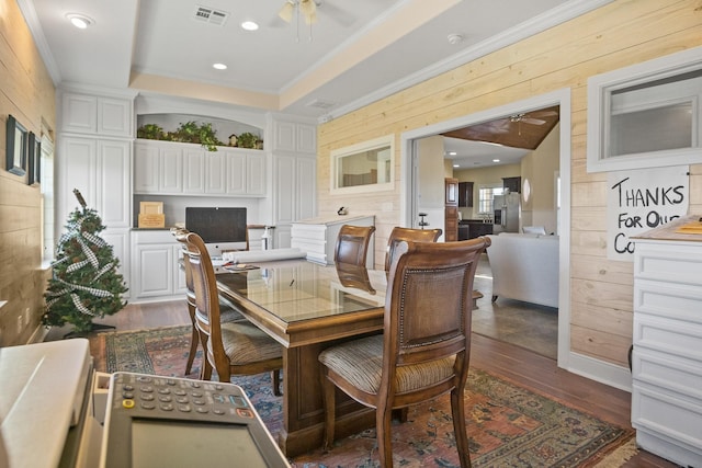 dining area with dark wood-type flooring, crown molding, a tray ceiling, wooden walls, and ceiling fan