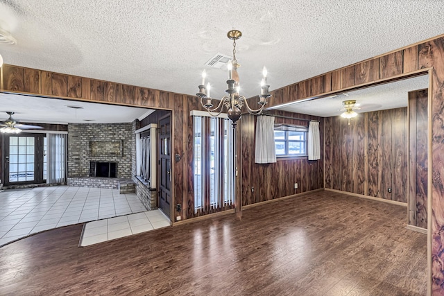 unfurnished dining area featuring ceiling fan with notable chandelier, a brick fireplace, wood walls, and a textured ceiling