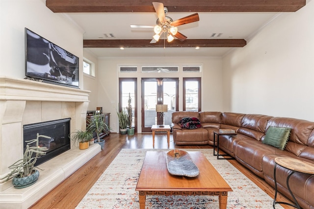 living room with wood-type flooring, crown molding, ceiling fan, a tile fireplace, and beamed ceiling