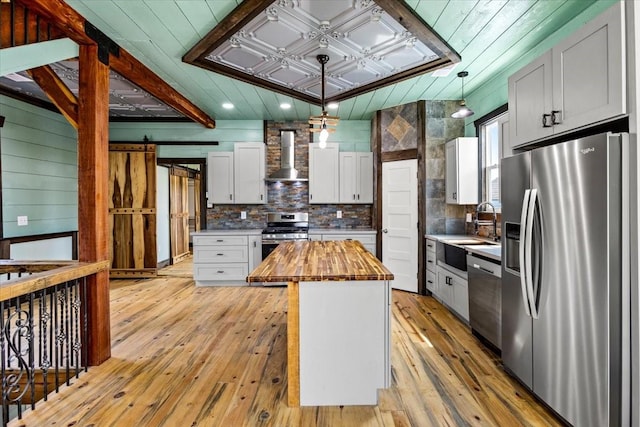 kitchen with pendant lighting, appliances with stainless steel finishes, wooden counters, a kitchen island, and wall chimney range hood