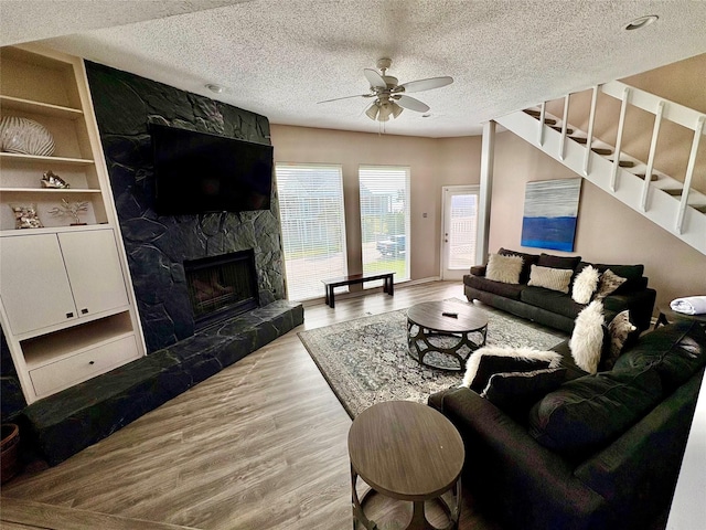 living room featuring a stone fireplace, a textured ceiling, light wood-type flooring, built in shelves, and ceiling fan