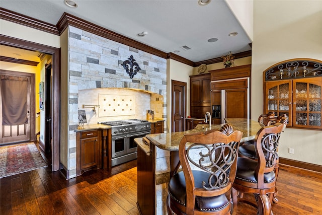 kitchen with crown molding, high end appliances, dark hardwood / wood-style flooring, light stone counters, and a center island with sink