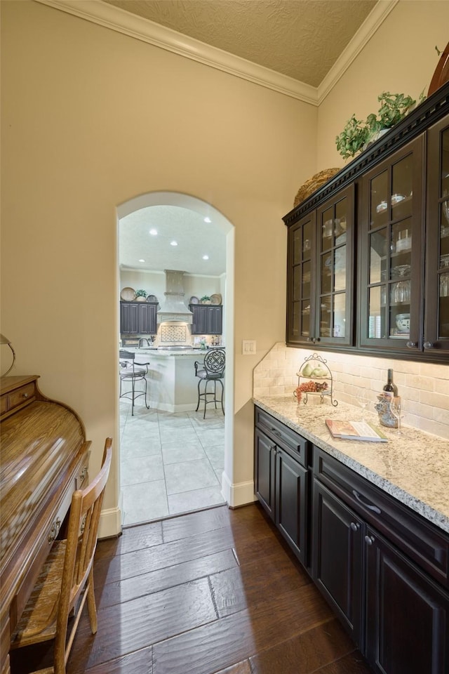 bar with decorative backsplash, custom exhaust hood, ornamental molding, light stone countertops, and dark wood-type flooring